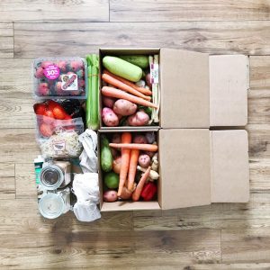 flat lay two cardboard boxes mixed vegetables in them, a plastic carton of 30% off strawberries, bag of oranges, bag of bean sprouts, and three cotton bags, two glass jars full of dry goods. All laying on a laminate floor.