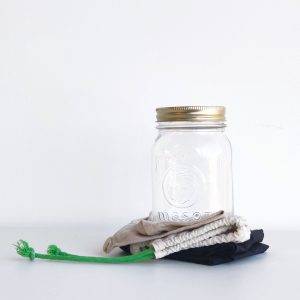 Zero Waste Shop Winnipeg - white background with a black nylon tote bag folded up under a cotton drawstring bag with green drawstring folded up with a glass mason jar atop both. Lid is gold.