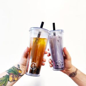 Zero Waste Shop Winnipeg - BYOC: Bubble Tea Edition two white arms coming in from each side of the frame holding clear plastic 750ml doubled walled cups with black straws. One cup full of passion fruit bubble tea and the other taro bubble tea. Both arms have tattoos. Background white.